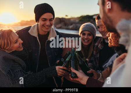 Group of multi-ethnic young people toasting beers on the beach. Friends spending time together at the beach with few drinks during sunset. Stock Photo