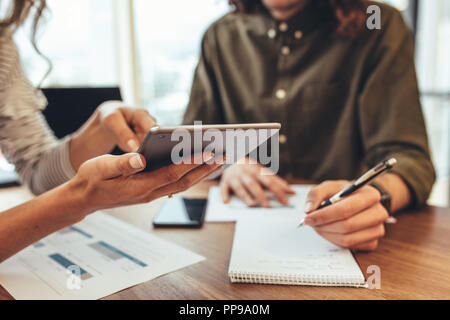 Woman showing a tablet computer to her colleague while discussing work in office. Man writing notes in book sitting at his work desk. Stock Photo
