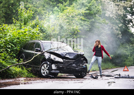 An angry young woman with smartphone by the damaged car after a car accident. Stock Photo