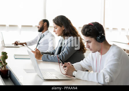 Portrait of three young people sitting in row and using gadgets at work in open office, copy space Stock Photo