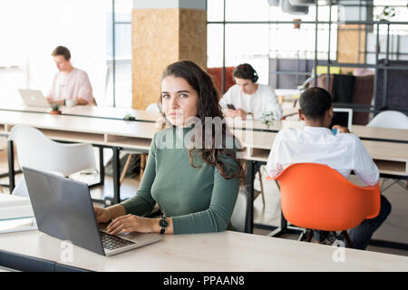 Portrait of beautiful young woman looking at camera while working with laptop in modem open office, copy space Stock Photo