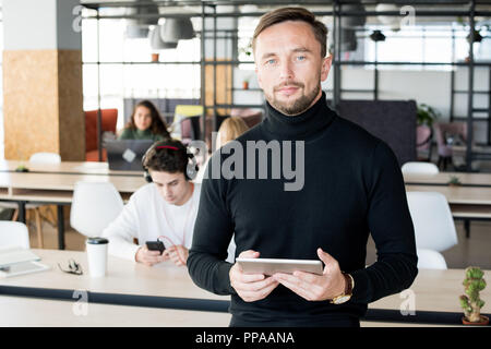 Waist up portrait of handsome contemporary businessman looking at camera and holding digital tablet while posing in open space office of startup compa Stock Photo