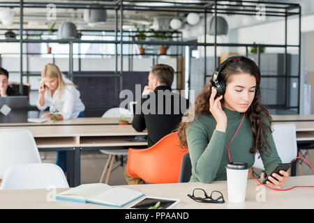 Portrait of contemporary young woman listening to music at workplace wearing big headphones in open space office Stock Photo