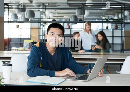 Portrait of young Asian man smiling at camera while working as intern in open space office of IT company Stock Photo