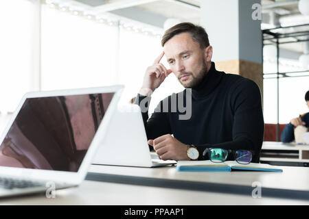 Portrait of handsome young man looking at laptop screen pensively while working in open space office Stock Photo