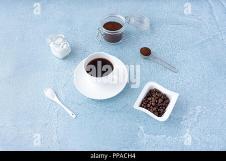 Coffee cup filled with black coffee, sugar, ground coffee and coffee beans on blue concrete background. Stock Photo
