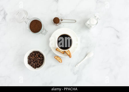 Black coffee in a cup with cantucci, white sugar, ground coffee and coffee beans on white marble background. Stock Photo