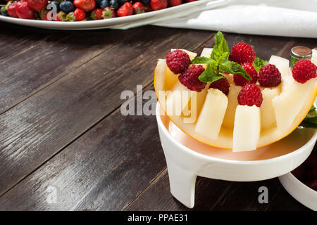 Raspberries lying on chopped melon on top mint lies in a white deep bowl on a brown wooden table in the background white tablecloth and a plate with b Stock Photo