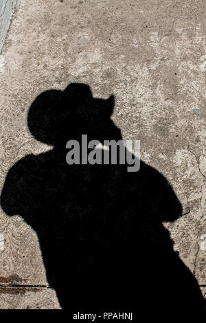 Image of a deep black shadow of a man's silhouette with a hat on brown and black spotted pavement Stock Photo