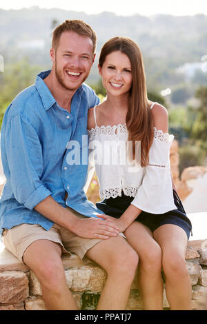 Portrait Of Romantic Young Holiday Couple Sitting On Wall Stock Photo