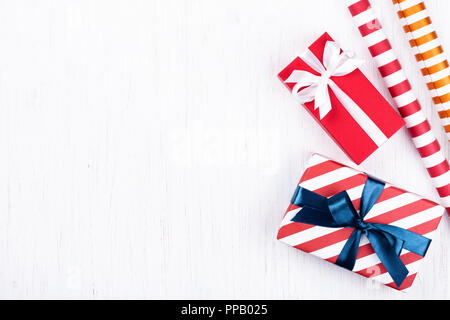 Christmas gift box wrapped in decorative striped and red paper tied with blue and white ribbons and two decorative paper rolls on white wooden backgro Stock Photo