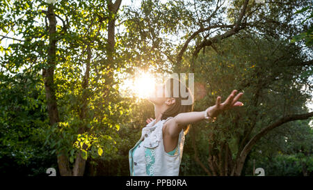Woman meditating in nature standing with outspread arms as the rising sun touches her face in a woodland setting , upper body in profile. Stock Photo