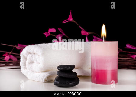 Aromatic candle burning on table and white towel and black stones, black background with purple flowers on branches. Spa treatment concept Stock Photo