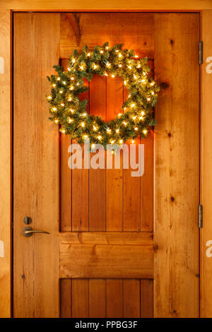 Beautiful lighted evergreen wreath hanging on wooden front door of home background. Simple, rustic country style Christmas holiday home decor. Stock Photo