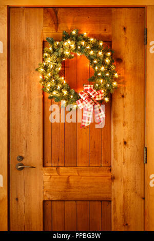 Beautiful Christmas decorations wreath with ribbon bow and lights on wooden front door background. Simple, rustic country style holiday home decor. Stock Photo
