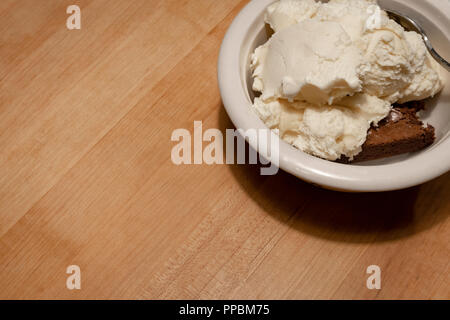 Ice cream and a spoon almost cover a brownie in a bowl on a wooden countertop. Stock Photo