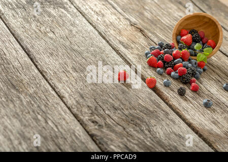 Delicious berry fruits, strawberries, blueberries, raspberries, blackberries, scattered from a wooden bowl on a textured rustic wooden desk with plent Stock Photo