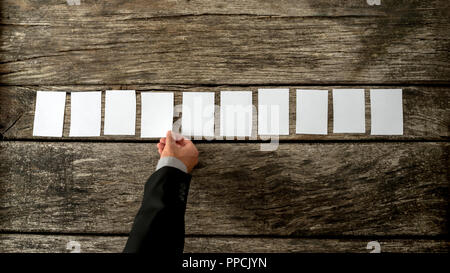 Overhead view of salesman placing 10 blank white cards in a row on a rustic textured wooden desk. Conceptual of marketing and sales, with copy space r Stock Photo