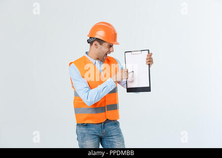 Frustrated young engineer with hardhat and reflective vest checking on mistake in document over gray background Stock Photo