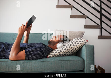 Man Lying On Sofa At Home Wearing Headphones And Watching Movie On Digital Tablet Stock Photo