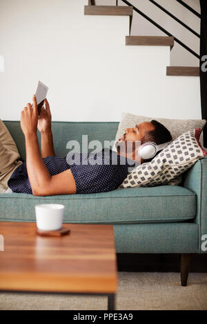 Man Lying On Sofa At Home Wearing Headphones And Watching Movie On Digital Tablet Stock Photo