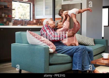 Grandparents Sitting On Sofa Playing With Baby Granddaughter At Home Stock Photo
