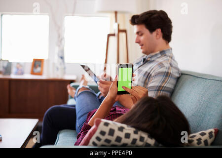 Couple Relaxing On Sofa At Home Using Mobile Phone And Digital Tablet Stock Photo