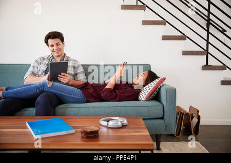 Couple Relaxing On Sofa At Home Using Mobile Phone And Digital Tablet Stock Photo