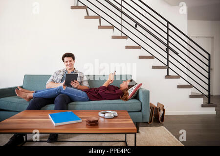 Couple Relaxing On Sofa At Home Using Mobile Phone And Digital Tablet Stock Photo