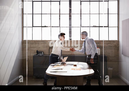 Two businessmen stand shaking hands in an office, side view Stock Photo