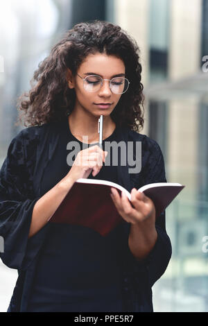 Charming Afro American beautiful happy female, making notes in notepad while walking outdoors on a city street. Education and business concept with sp Stock Photo
