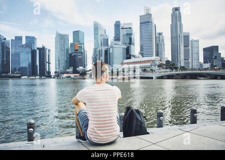 Young man (traveler) with backpack sitting on waterfront against downtown and financial district of Singapore. Stock Photo