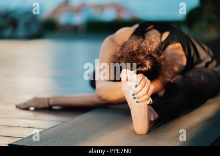 Stretching close up foot. Girl doing split twine. Hanumanasana, monkey pose. Young oriental appearance woman practicing yoga on wooden deck in tropica Stock Photo