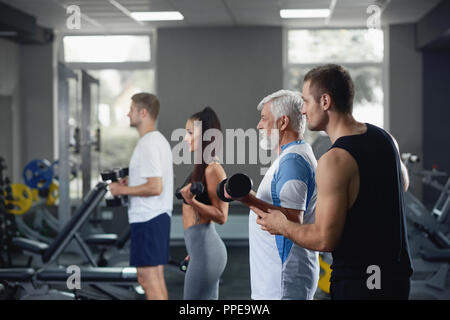 Elderly man doing exercise with dumbbell at gym with group of diverse  younger people who workout with same equipment. Team working together under  supervision of personal coach, training their muscles Stock Photo 
