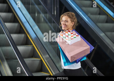 View from side of cheerful lady with shopping bags, looking at camera and laughing in mall. Attractive young woman driving up on escalator. Concept of enjoying shopping and happiness. Stock Photo