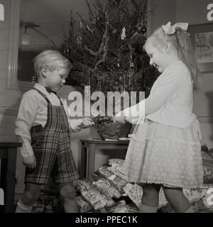1950s, historical, a young boy and girl pull a christmas cracker in front of a small decorated christmas tree and a with pile of presents underneath, England, UK. The traditional of pulling crackers is said to date back to the mid 1850s when they were first made by a man called Tom Smith, who got the idea from the 'crackle' an open fire makes. Stock Photo