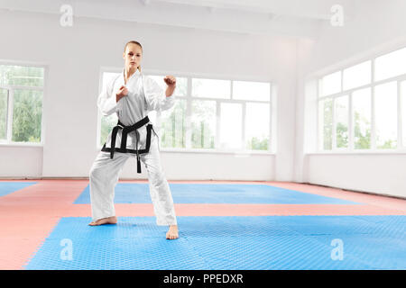 Professional female fighter standing in pose with clenched fists, ready for battle. Blonde girl wearing in white kimono with black belt practicing martial arts at karate class. Concept of sport. Stock Photo