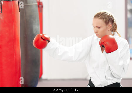 Seriously pretty woman hitting boxing heavy bag in gym. Sporty karate girl, wearing in kimono and red protective gloves improving technique of fight at hard mixed martial arts training. Stock Photo