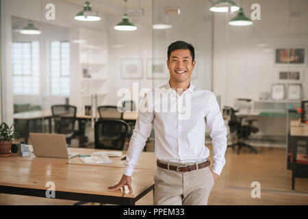 Young Asian businessman standing in an office smiling confidently Stock Photo