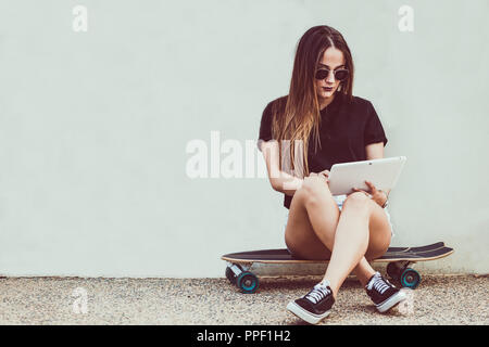 Young woman sitting on skateboard and holding tablet Stock Photo
