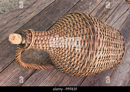 Old demijohn wicker wrapped glass bottle on wooden table Stock Photo