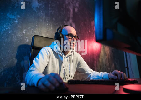 Portrait of a joyful young gamer playing video games at home on a computer. Gamer in headphones is happy and showing thumbs up gesture because he has won the game. Computer Games Concept. Stock Photo