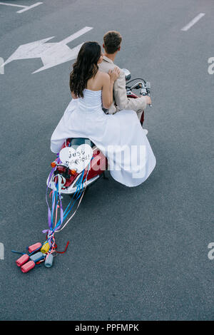 back view of wedding couple riding scooter with 'just married' heart sign and colorful cans on ribbons Stock Photo