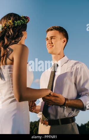 low angle view of happy young wedding couple holding hands and smiling each other against blue sky Stock Photo