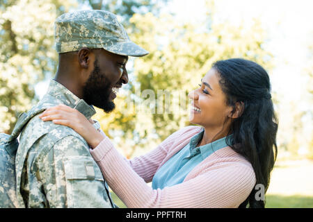 side view of smiling african american soldier in military uniform hugging girlfriend in park Stock Photo