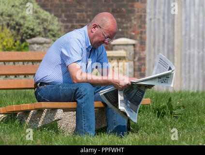 Smart casually dressed caucasian middle aged man sitting on a wooden park bench reading a newspaper in the UK. Stock Photo