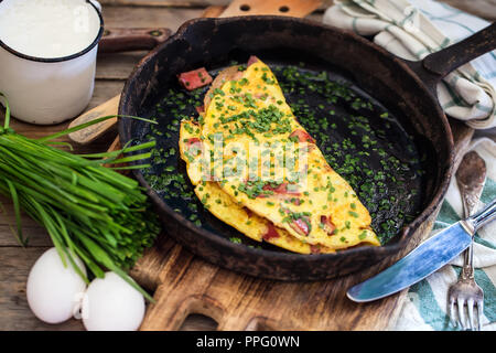 Cast iron frypan with omlette with ham and chives on old wooden background. Rustic style. Stock Photo