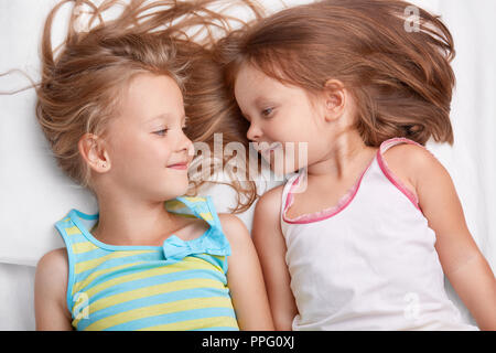 Two small sisters sleep together, look with positive expressions at each other, dressed in casual clothes, have good relationships. Best kid female fr Stock Photo