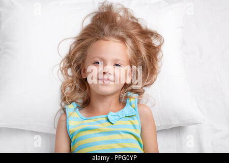 Attractive small kid with pleasant appearance, blue eyes and healthy skin looks at camera, poses in bed at bedroom, has refreshed look after having lo Stock Photo
