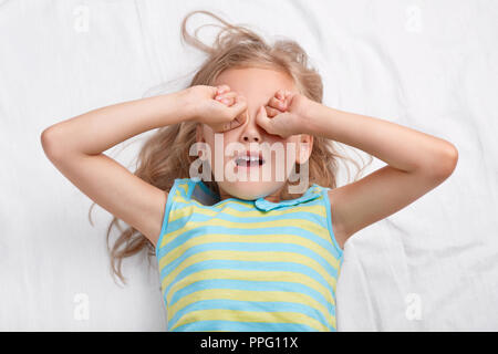 Photo of pleasant looking light haired girl rubs eyes, wants to sleep, dressed in nightwear, lies on comfortable bed, needs rest. Small relaxed sleepy Stock Photo